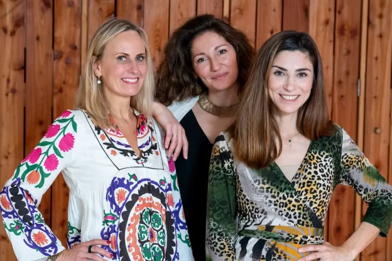 Three of Rejuvyn's expert facilitators smile and pose together in colorful patterned dresses, standing in front of a wooden wall.