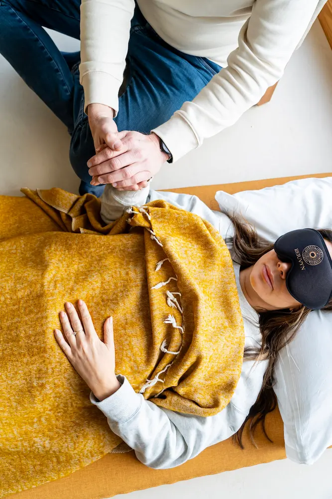 A person lies on a bed covered with a yellow blanket, wearing an eye mask during a psilocybin session, while another person sits beside them, gently holding their hand for comfort.