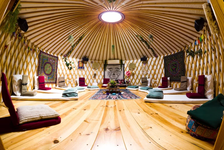 The interior of a cozy yurt features a wooden floor, round skylight, floor cushions with blankets, low tables, and colorful tapestries hanging on the lattice walls. The yurt’s space feels inviting and peaceful.