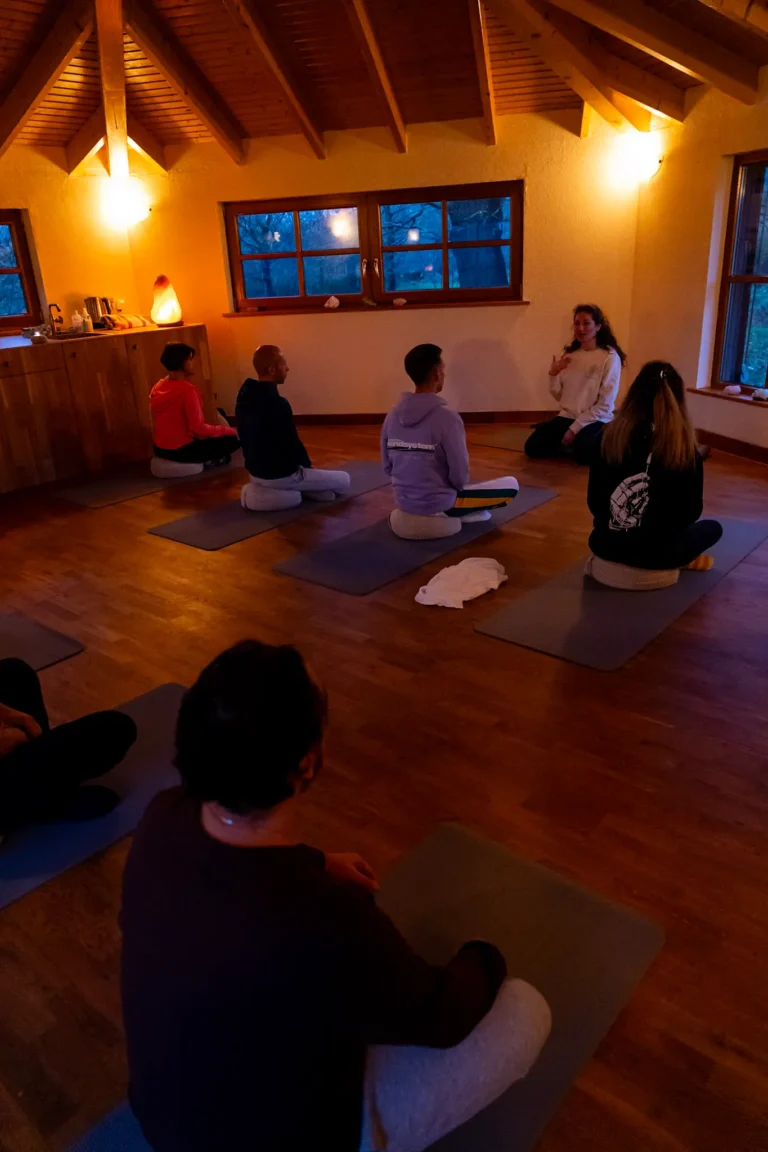A small group of people sits on yoga mats in a cozy, softly lit room with wooden floors and ceiling, attentively listening to an instructor at the front.