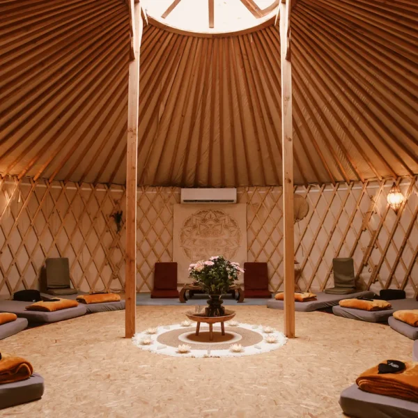 Interior of a cozy yurt with a circular skylight, wooden beams, cushions in a circle on the floor, colorful rugs, plants, and decorative wall hangings, creating the warm, inviting atmosphere typical of a traditional yurt.